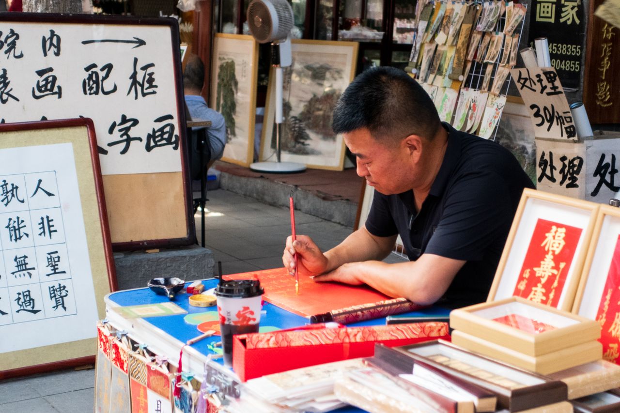 An Asian man focuses on traditional calligraphy art in an outdoor market setting.